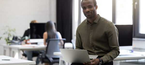 Homme d'affaires afro-américain souriant et heureux assis sur son bureau et tenant un ordinateur portable dans un bureau